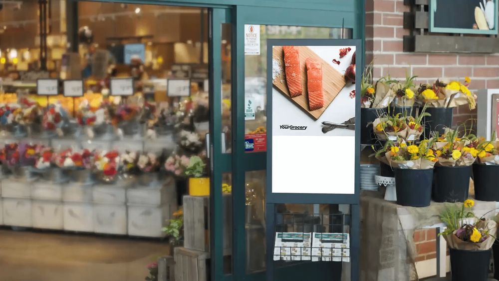 A digital screen at a supermarket entrance, beside the flower display.